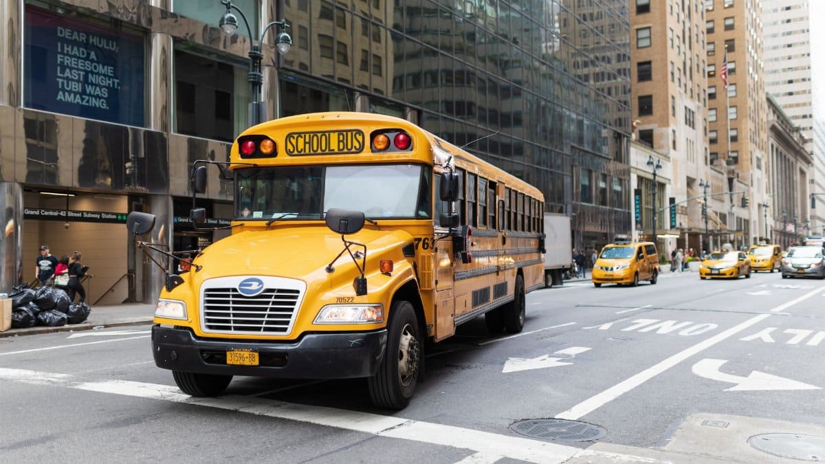 A yellow school bus navigates a busy New York City street amidst tall buildings and taxis.