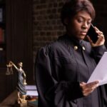 African American judge in robe, reading document and talking on phone by the office window.