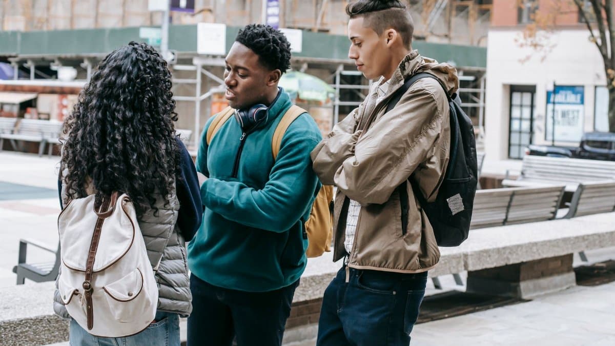 Multiethnic classmates in casual outfits and backpacks standing on city street near buildings and benches while having argument
