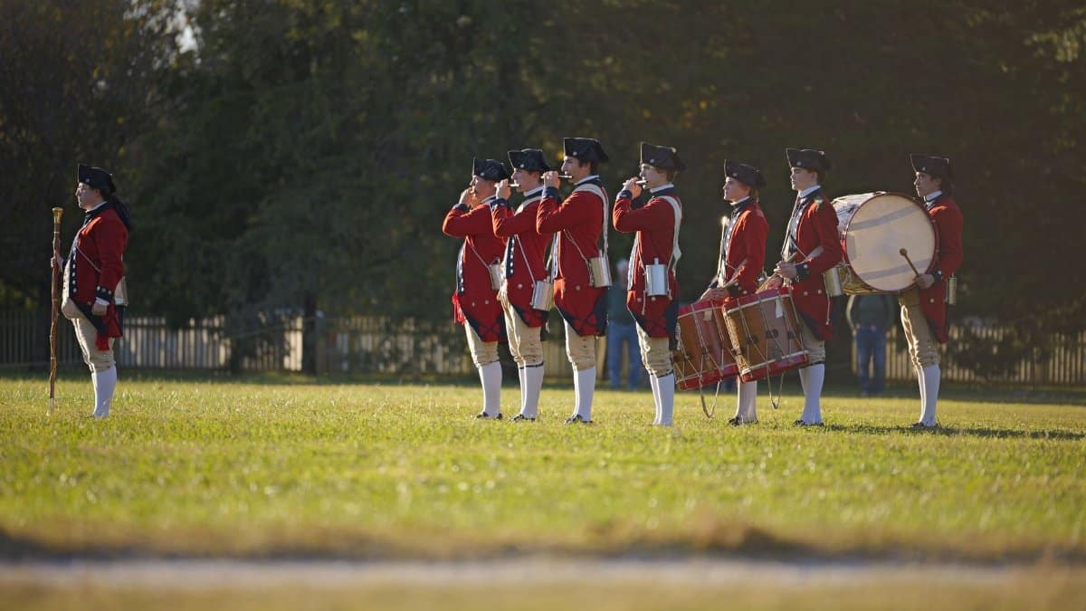 Colonial-era military band reenacting history in open field.