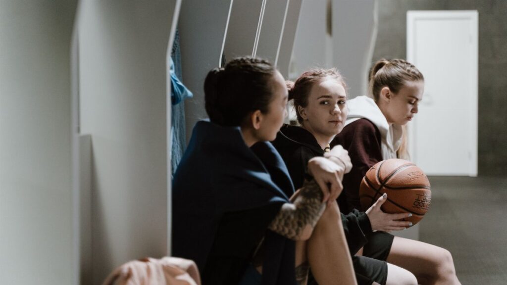 Three teenage girls sitting in a locker room, holding a basketball and chatting after practice.