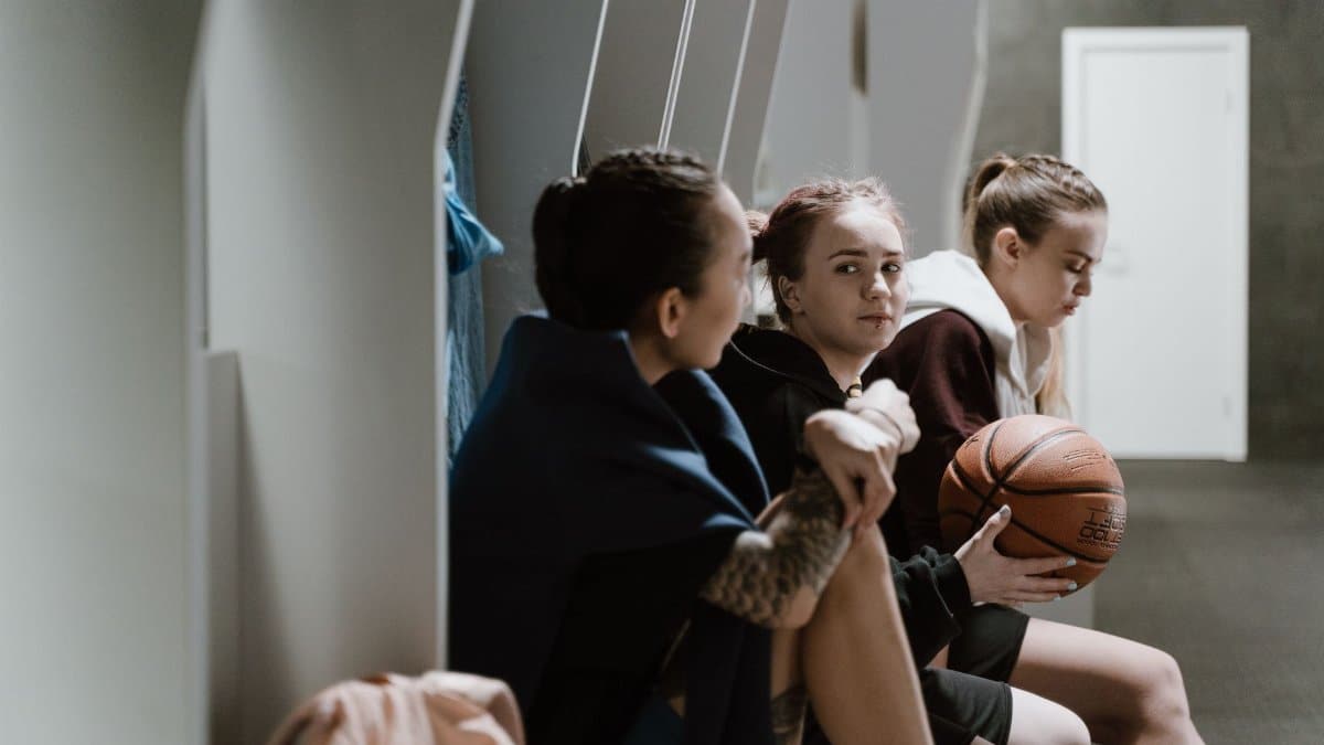 Three teenage girls sitting in a locker room, holding a basketball and chatting after practice.