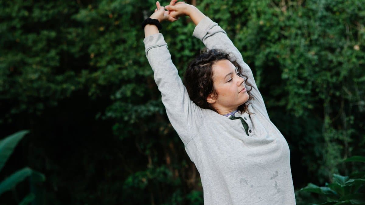 Calm young female with long curly hair in casual clothes warming up arms with closed eyes before outdoor training in green forest