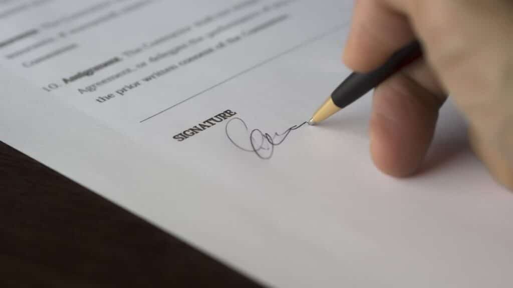 A hand signs a formal contract with a pen on a wooden desk.