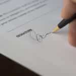 A hand signs a formal contract with a pen on a wooden desk.