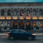 A dynamic street view of the Boston Public Library at dusk featuring American flags and illuminated decorations.