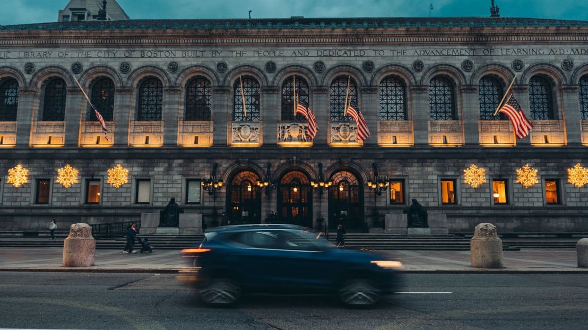 A dynamic street view of the Boston Public Library at dusk featuring American flags and illuminated decorations.