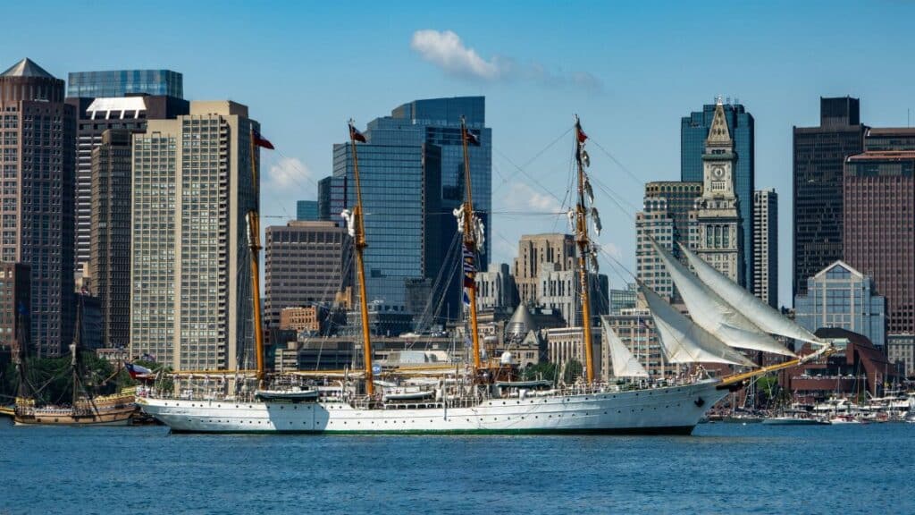 A majestic tall ship sails past the iconic Boston skyline on a clear summer day.