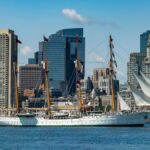 A majestic tall ship sails past the iconic Boston skyline on a clear summer day.