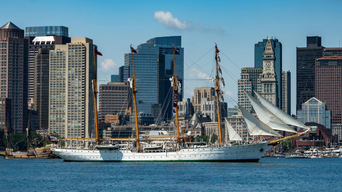 A majestic tall ship sails past the iconic Boston skyline on a clear summer day.