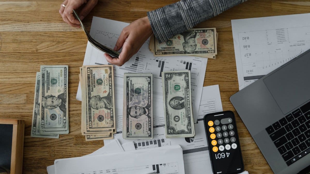 Person counting cash with a calculator and documents on a wooden desk, representing financial planning.