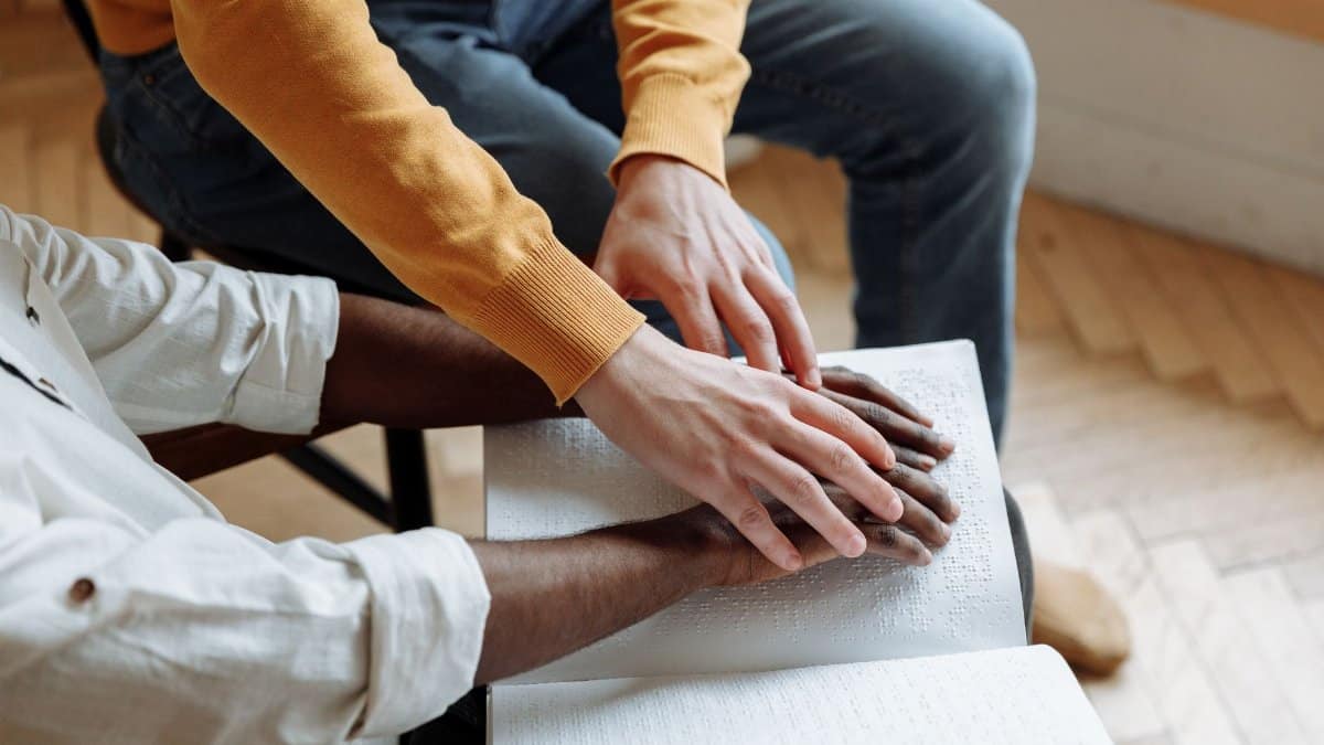 Man guiding another in reading braille indoors, illustrating support and learning.