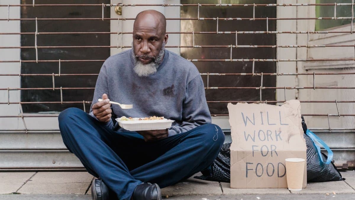 Elderly homeless man sitting on street with a sign and meal, symbolizing poverty.