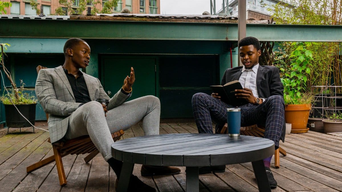 Two men in suits having a conversation outdoors, one reading a book.