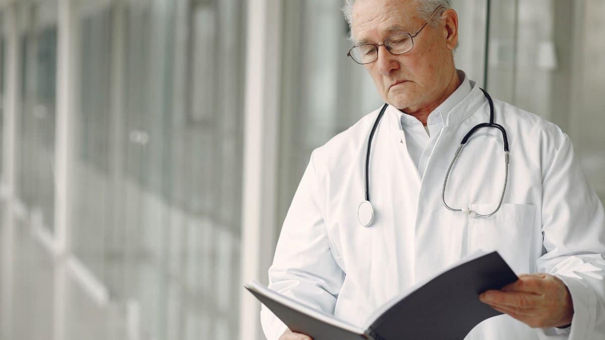 Crop concentrated professional senior male physician in white robe with stethoscope standing in modern hospital hallway and reading medical case records in daylight
