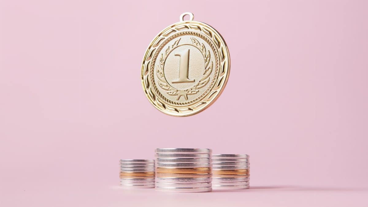 A gold medal and stacks of coins on a light pink backdrop, symbolizing victory and wealth.