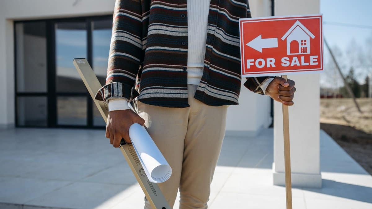 A real estate agent stands outside with a 'for sale' sign and rolled blueprint, suggesting a property for sale.