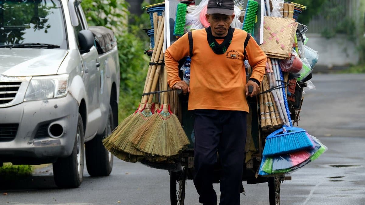 A man in an orange shirt pulls a cart loaded with household goods on a street during the day.