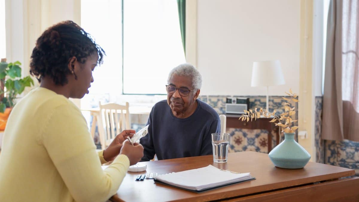 A caregiver assists an elderly man with medication in a warm, sunlit living room.