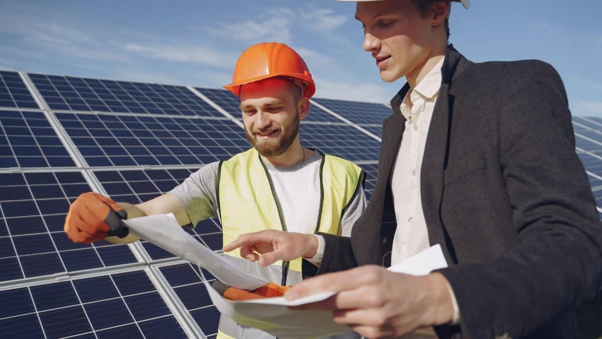 Low angle of young engineer and businessman in hardhats discussing project details against solar energy station