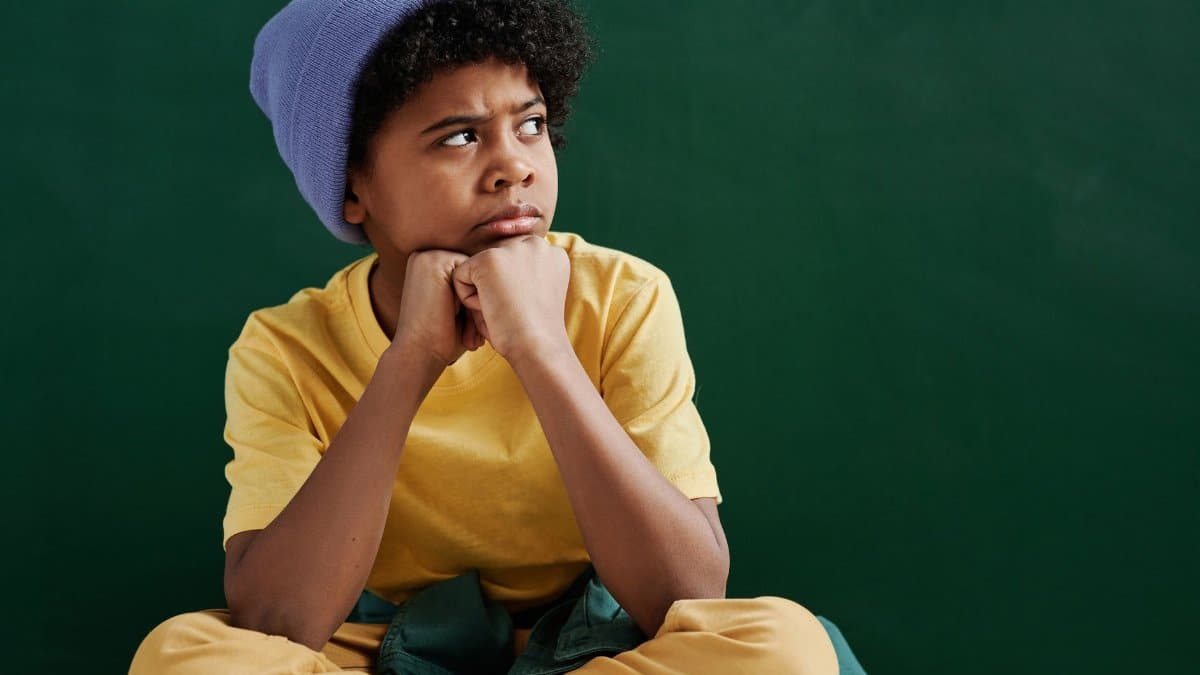 Pensive boy with curly hair wearing a yellow shirt and blue beanie against a green background.