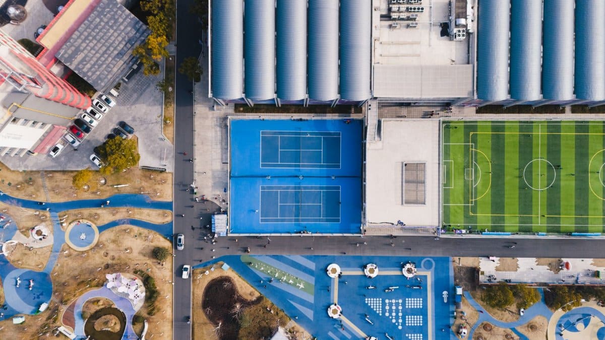 Aerial shot of a sports complex showing tennis courts, soccer field, and colorful playground.