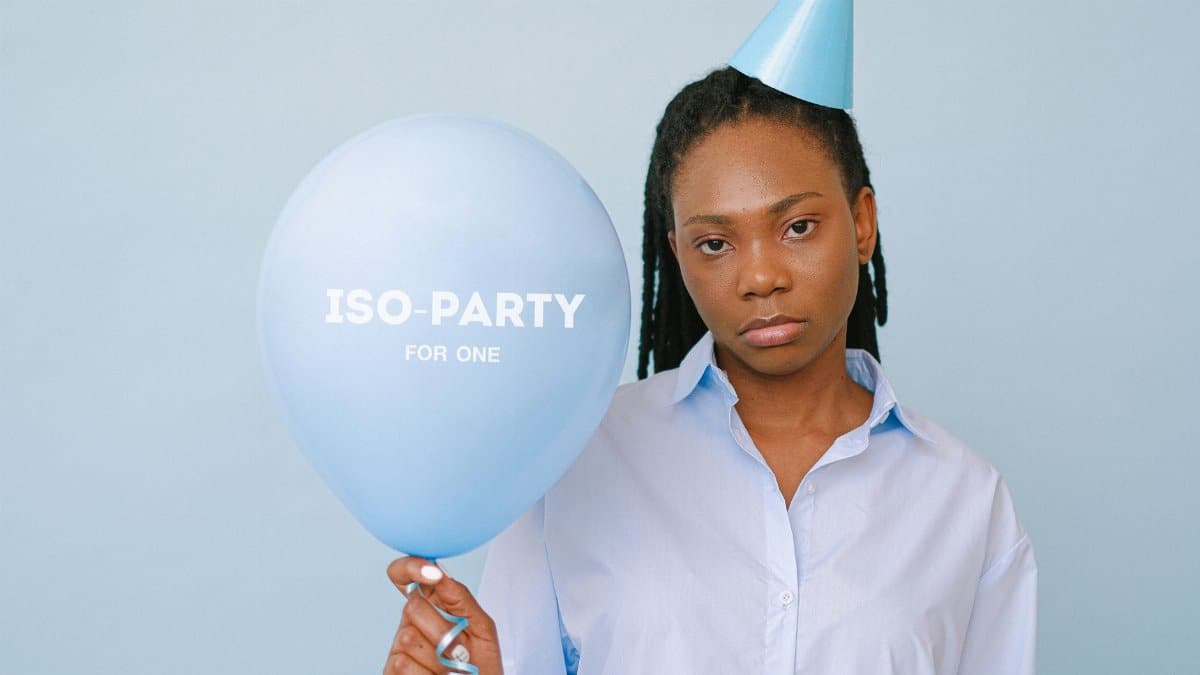 A somber woman holding a balloon with 'ISO-Party for One' text, wearing a party hat.