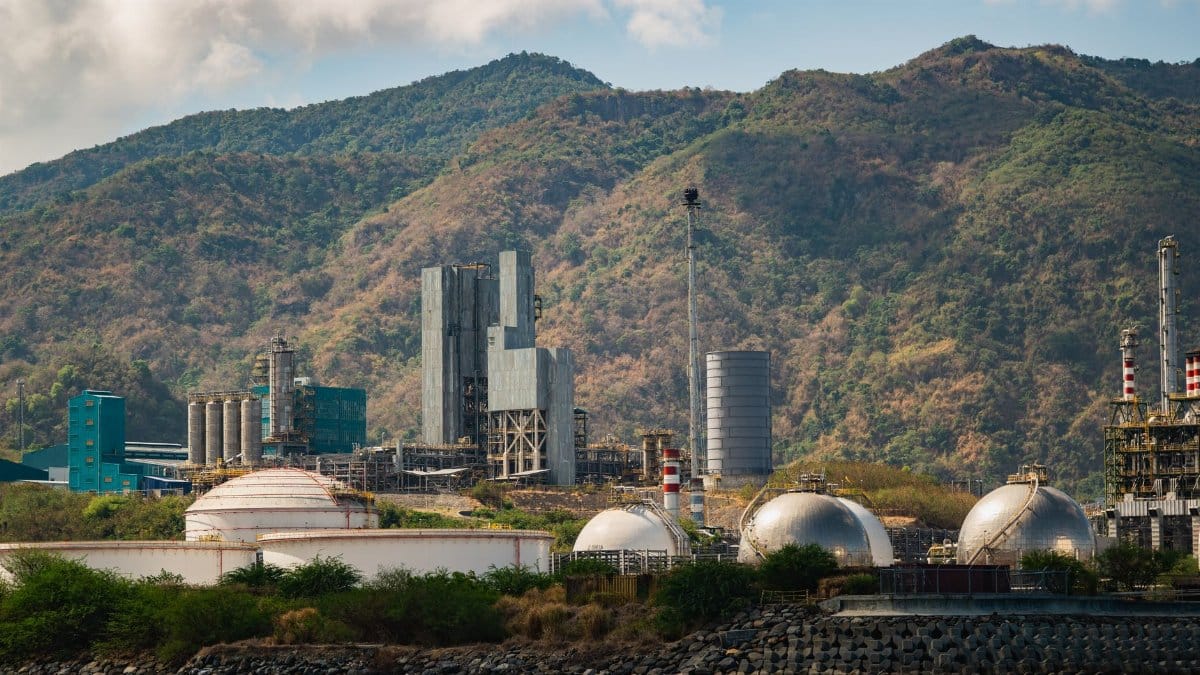 Industrial gas tanks and chimneys against lush hills, showcasing industry and nature.