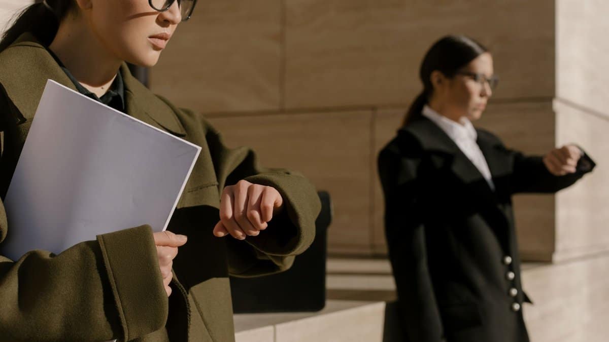 Two businesswomen wearing coats, checking time in bright daylight.