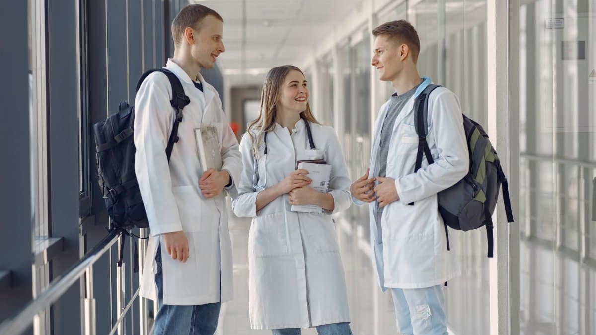 Three young medical professionals in lab coats talking in a hospital hallway.