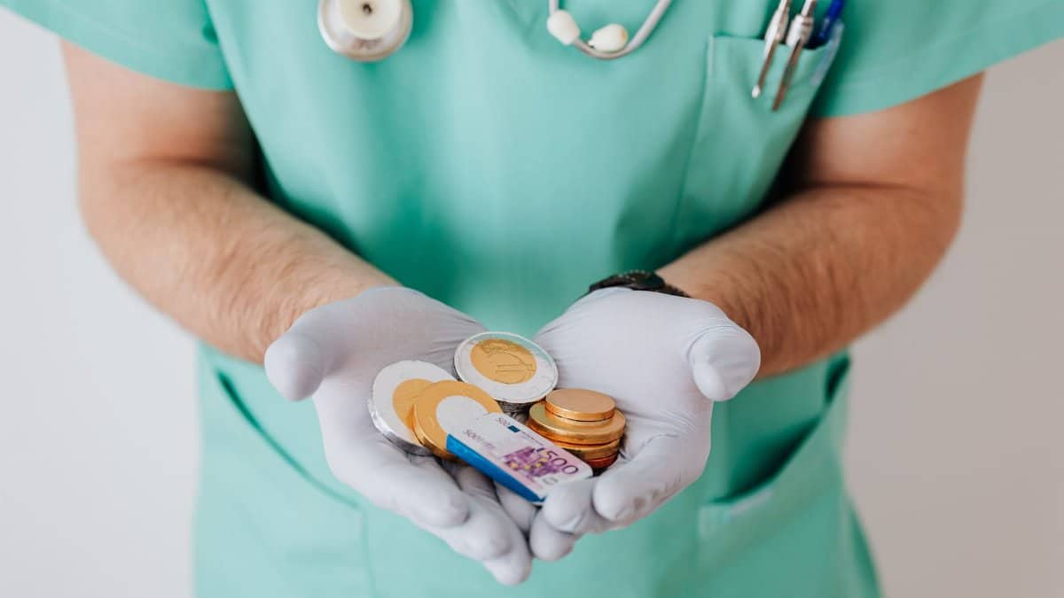 Unrecognizable medical worker demonstrating money wearing medicine form standing near white wall in hospital while working in day of salary