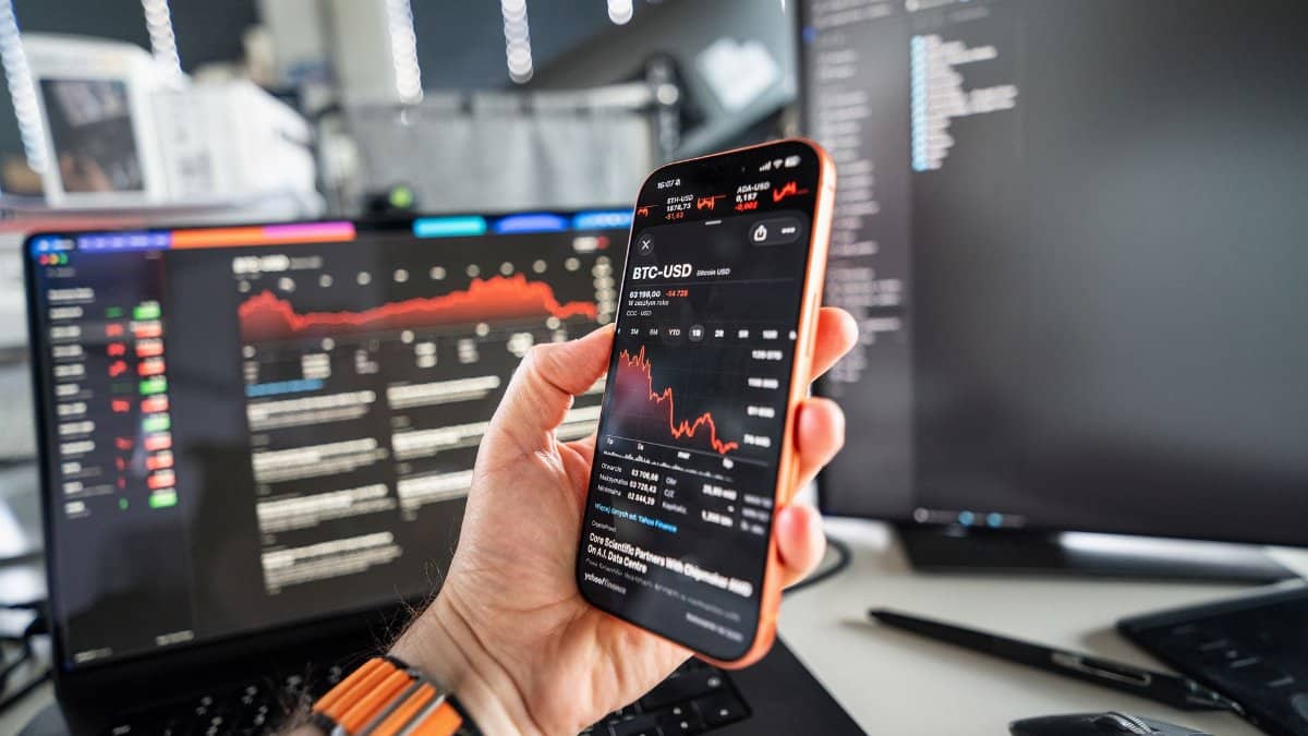 A person analyzing cryptocurrency trends on a smartphone with a declining chart on screen, set against a tech office background.