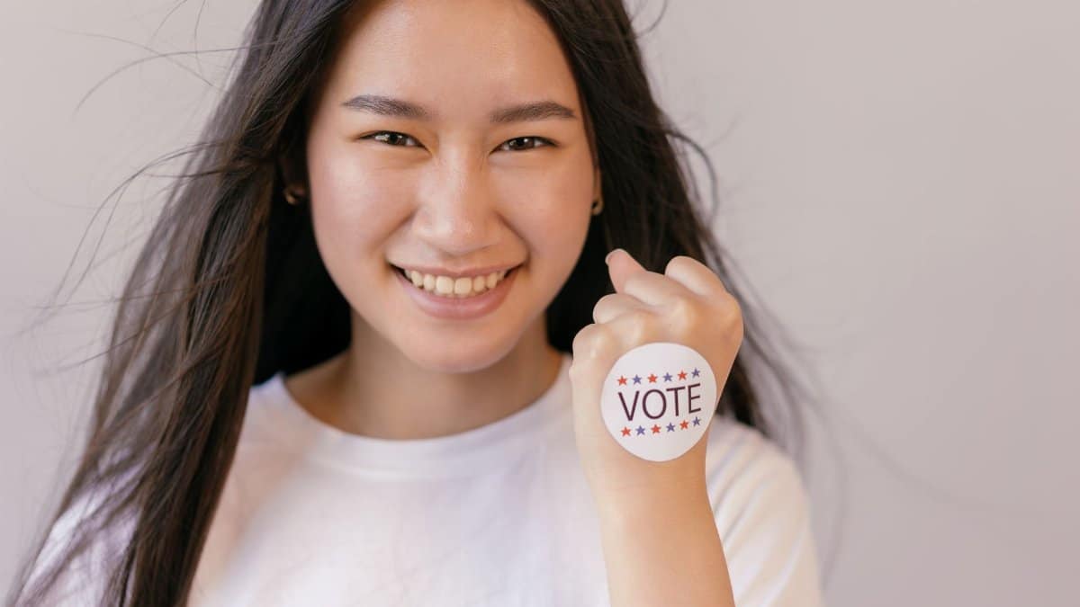A smiling young woman holds up a vote sticker, promoting the spirit of democracy and participation.