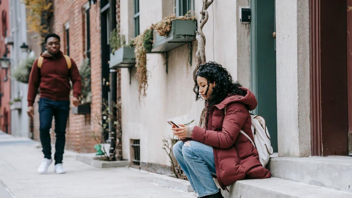 Young female using mobile phone and resting on gray steps of building while African American male promenading on street