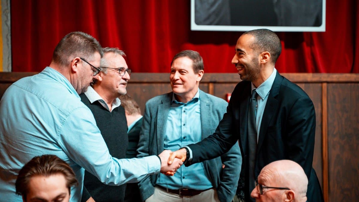 Five men in formal attire shaking hands and conversing at an indoor business meeting.