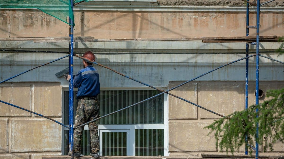 A construction worker in a hard hat repairs a building facade on scaffolding in an urban setting.