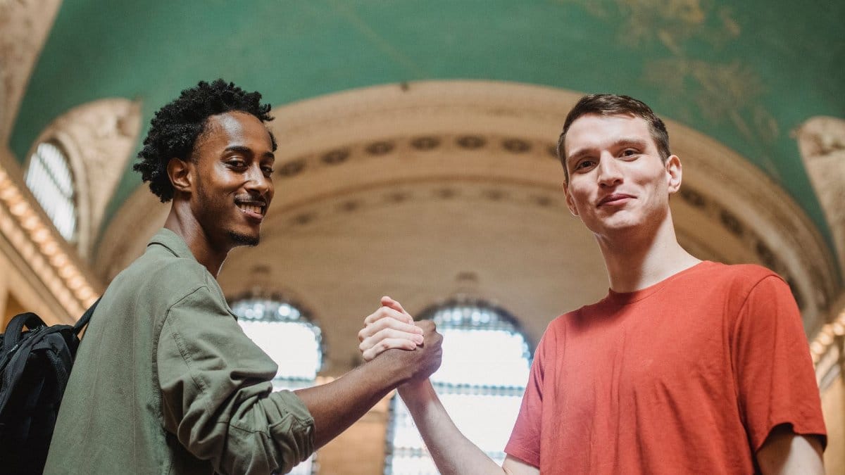 From below of positive young multiethnic male friends in casual clothes smiling and looking at camera while shaking hands in spacious aged arched building