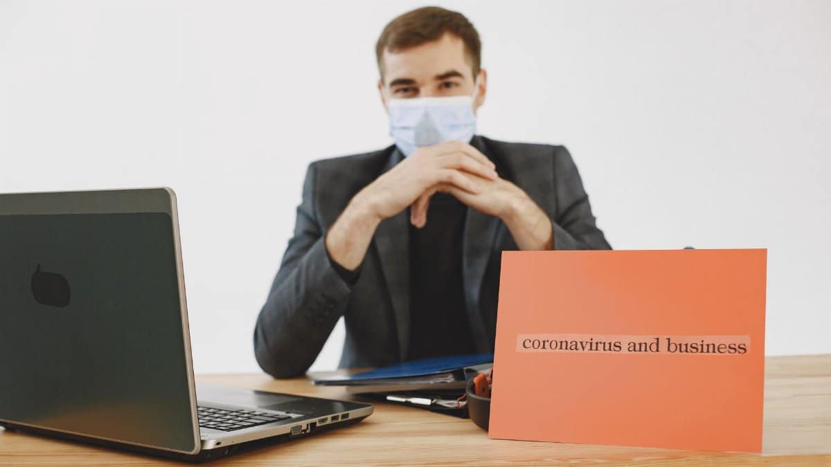 Man in office wearing face mask, working on laptop with sign about coronavirus and business.