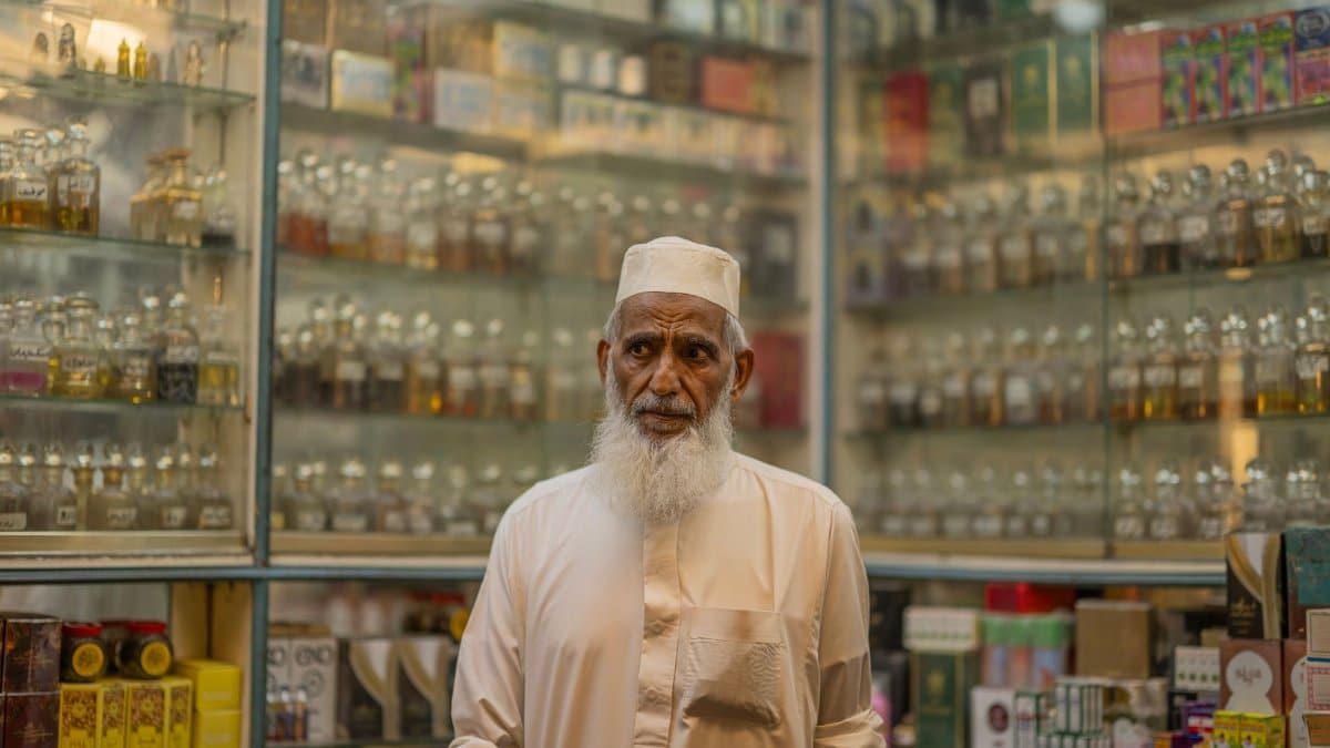 Elderly man wearing traditional clothing in an aromatic perfume shop with glass shelves.