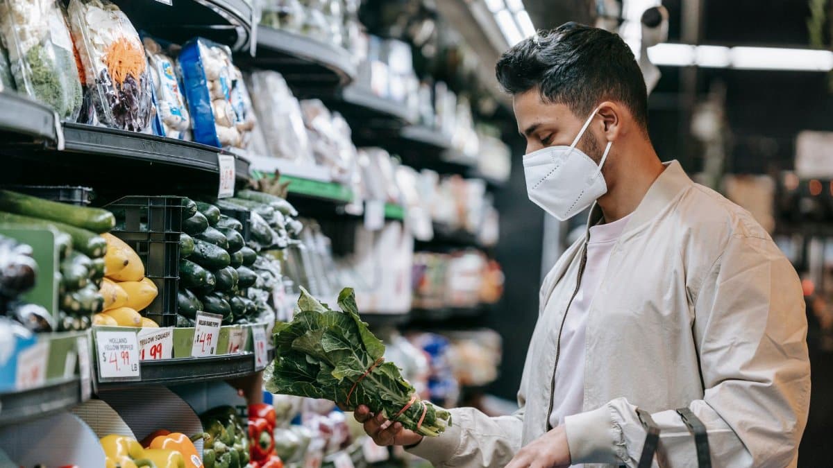 Side view of young ethnic male buyer in trendy jacket and medical mask choosing spinach during shopping in modern hypermarket