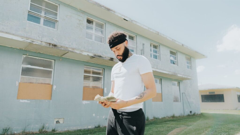 A young man with facial hair counts money in front of a worn building on a sunny day.