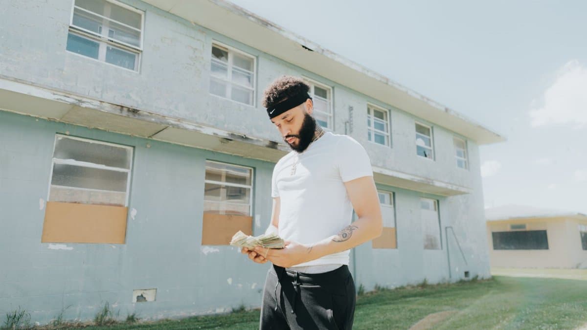 A young man with facial hair counts money in front of a worn building on a sunny day.