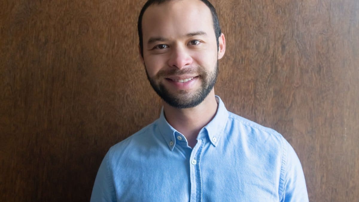 A portrait of a smiling man wearing a blue shirt standing against a wooden background indoors.