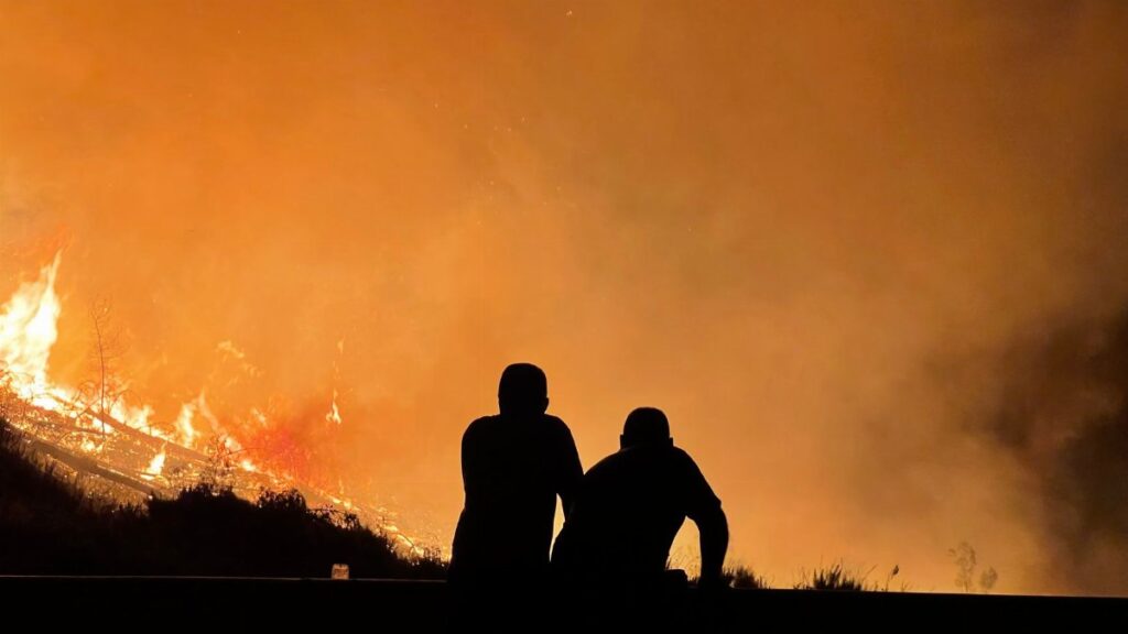 Silhouetted figures observe a large wildfire engulfing the landscape, filling the sky with orange flames and smoke.