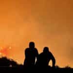 Silhouetted figures observe a large wildfire engulfing the landscape, filling the sky with orange flames and smoke.