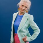 Delightful gray haired female in stylish green suit putting hand on waist and looking at camera against blue background