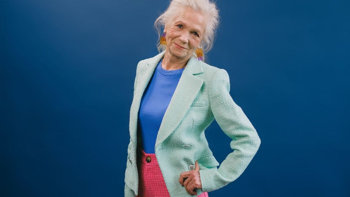 Delightful gray haired female in stylish green suit putting hand on waist and looking at camera against blue background