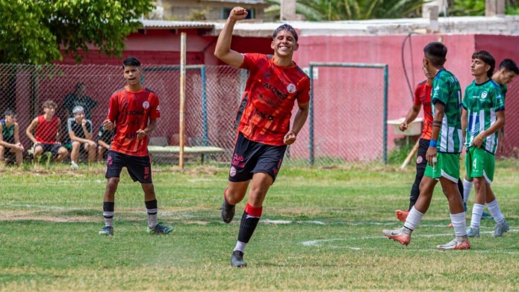 A teenage soccer player celebrates scoring a goal during a match. Teams in red and green jerseys.