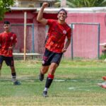 A teenage soccer player celebrates scoring a goal during a match. Teams in red and green jerseys.