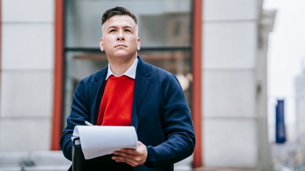 A thoughtful man in professional attire sitting outdoors, looking up pensively.